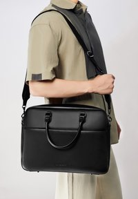Maletín de cuero negro con doble asa y correa desmontable. Textura suave, forma rectangular y diseño minimalista.