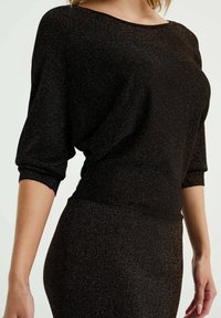 Svart, glittrig stickad topp med vid halsringning och trekvartslånga ärmar. Mjuk textur med avslappnad passform, rynkad vid midjan.