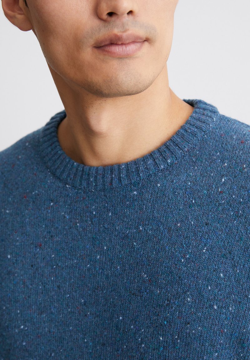 Blauer gestrickter Pullover mit rundem Ausschnitt, der einen strukturierten Stoff und mehrfarbige Flecken aufweist. Nahaufnahme des Oberkörpers.