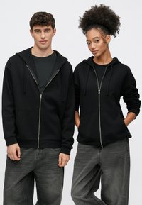 Felpe con cappuccio nere con cerniera, realizzate in tessuto morbido, con coulisse e tasche frontali, indossate sopra pantaloni grigi. Design semplice e casual.