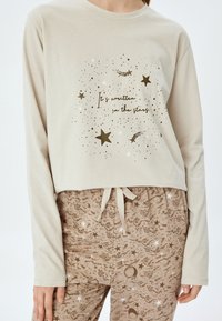 Beige langlopende top met een bedrukte afbeelding van sterren en tekst. Bruine pyjamabroek met zwierige patronen, sterren en manen. Koordsluiting in de taille.