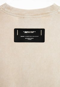 Camiseta de algodón beige con un escote acanalado que presenta una etiqueta de cuero negro con la inscripción "MWM" y texto sobre la marca y la agencia de diseño.
