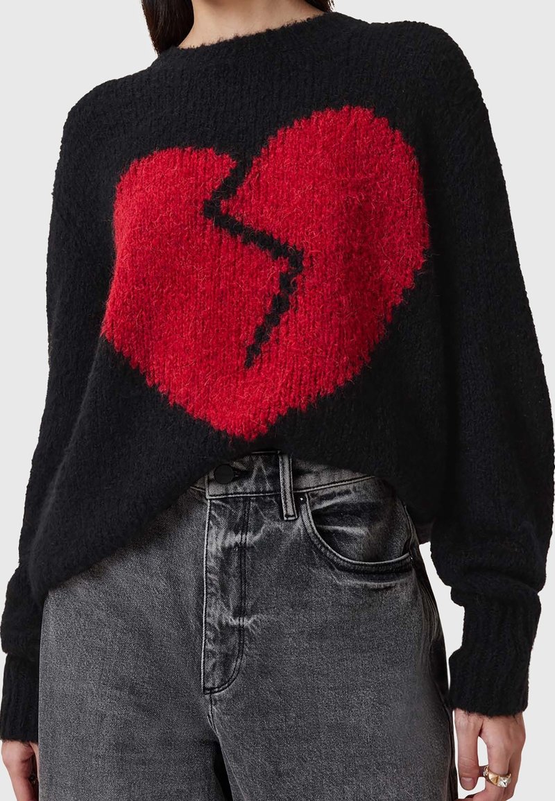 Czarny, teksturowany sweter z dużym czerwonym sercem o czarnym wzorze pęknięć. Noszony z szarymi spodniami z wysokim stanem. Długie rękawy.