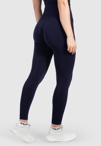 Leggings blu navy, realizzati in tessuto elastico, a vita alta con una texture liscia e cuciture laterali. Abbinati a scarpe da ginnastica bianche.