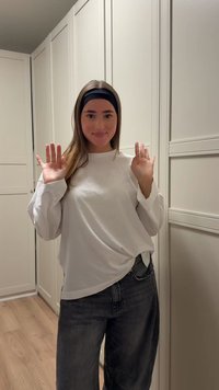 White long-sleeve shirt with loose fit, paired with dark gray baggy jeans. Hands raised, in a light-colored room with wooden flooring.