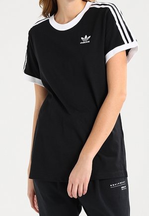 Kobieta w czarnej koszulce Adidas z białymi lamówkami i trzema paskami na rękawach, w połączeniu z czarnymi spodniami.