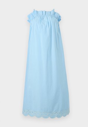 Ψηλή γυναίκα με αμάνικο μεσομήκες φόρεμα γαλάζιου χρώματος με βολάν στο ντεκολτέ, κρατώντας μικρή απαλό ροζ τσάντα, φορώντας μαύρα φλατ παπούτσια, στέκεται μπροστά σε λευκό φόντο.