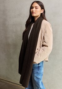 Doudoune beige avec un design court, associée à une écharpe en côtes marron foncé. Un jean en denim clair complète la tenue.