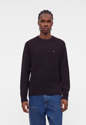 Jeune homme aux cheveux tressés portant un pull à manches longues violet foncé et un jean bleu, debout devant un fond clair uni.