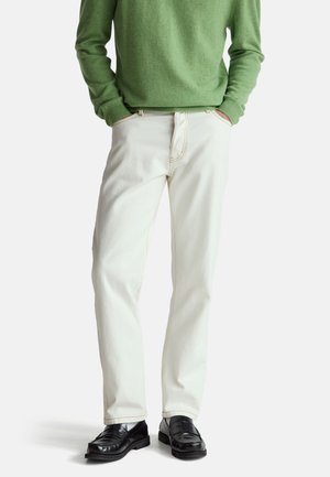 Mann trägt hellbeige Hose, schwarze Loafers und einen grünen Langarmpullover, die Hände in den Taschen.