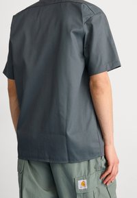 Personne portant un t-shirt gris foncé à manches courtes et un pantalon cargo vert clair avec un patch logo Carhartt sur la poche, vue de dos.