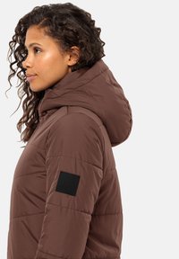 Braune Daunenjacke mit Kapuze, die ein gesteppte Design aufweist und ein schwarzes Logo-Patch am linken Arm hat. Glatte Textur, Seitenansicht.