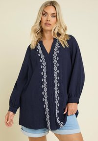 Camicia tunica blu navy con motivi ricamati bianchi lungo la parte anteriore e tessuto morbido e textured. Maniche lunghe con polsini elasticizzati.