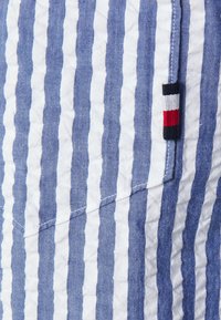 Puuvillaiset shortsit, joissa on vaakasuorat sinivalkoiset raidat. Sivulla on pieni, kolmivärinen logopainatus ja kankaassa on tekstuuri.