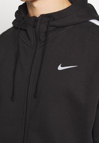 Svart zip-up huvtröja med hög krage, som har dragsko och en vit Nike-logotyp på bröstet. Slät textur och sportig design.