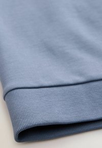 Modrý manžetový svetr, který se vyznačuje žebrovanou texturou, hladkým materiálem a mírným elastickým účinkem. Design zdůrazňuje čisté provedení a jemný lesk.