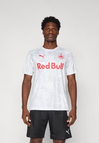 Grå camouflagemønstret sportsjersey med "Red Bull" i fed rød tekst, Puma-logo i rød, båret med sorte atletiske shorts.