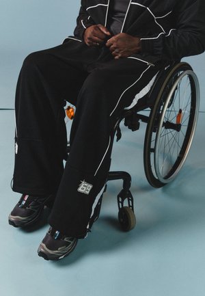 Black athletic pants with white side stripes, worn with black shoes featuring gray accents. Model seated in a wheelchair on a light blue background.