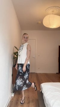 Bílý bezrukávový top s mašlí, zkombinovaný s volnou černou sukní s modrým květinovým vzorem. Černé flip-flopy. Dřevěná podlaha.