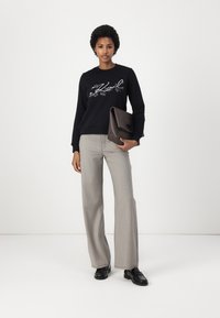 Sudadera negra con gráfico detallado en blanco, pantalones acampanados gris claro y un clutch marrón oscuro; la modelo lleva mocasines negros.