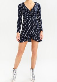 Námořnické zavinovací šaty s dlouhými rukávy, bílými puntíky, volánkovým výstřihem a rozšířeným lemem. Kombinováno s metalickými stříbrnými kotníkovými botami.