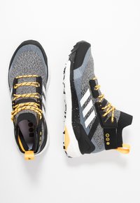 Chaussures de sport noires et grises avec une tige texturée, des accents jaunes, trois bandes blanches et un col semi-haut ; dispose d'une semelle extérieure noire.