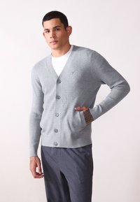 Šedý pletený cardigan s výstřihem do V, zapínáním na knoflíky a žebrovanými manžetami. Má kapsu na levé straně a hladký povrch.