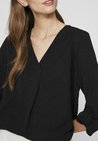 Svart, texturerad blus med v-ringning, trekvartslånga ärmar och uppvikta manschetter. Har en draperad design som förbättrar komfort och passform.