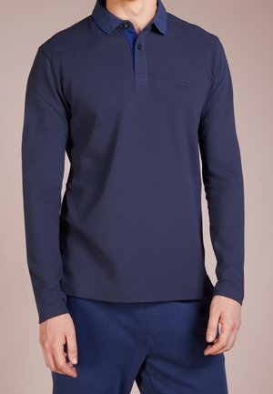 Mann trägt ein marineblaues Langarm-Poloshirt und passende Hosen, steht mit entspannten Händen an den Seiten vor einem schlichten Hintergrund.