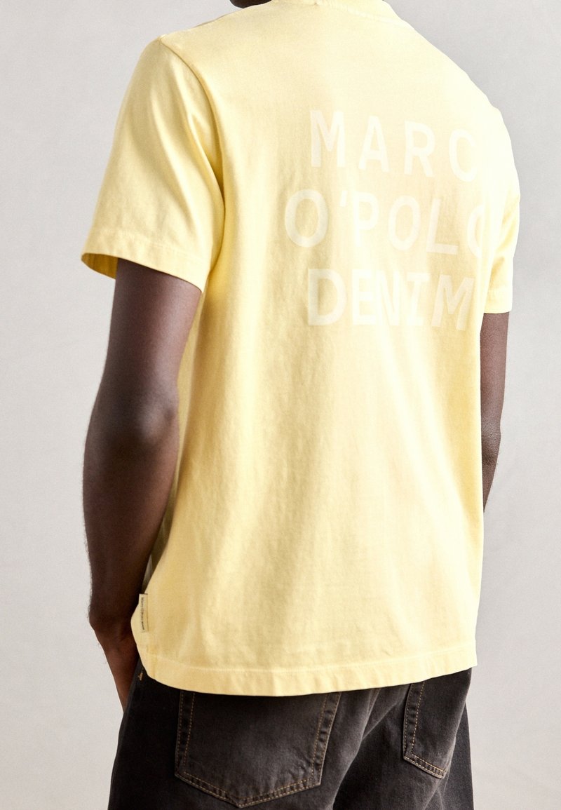 Camiseta de algodón amarilla con cuello redondo y mangas cortas. Presenta un texto grande y de color claro en la parte posterior que dice "MARC O'POLO DENIM."