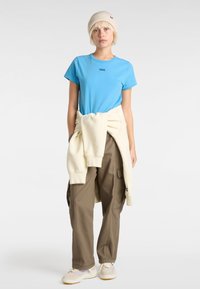 Sininen puuvillapaita, jossa on Vans-logo, kerma väri neule vyötärölle sidottuna, oliivinvihreät cargo-housut ja vaaleita yksityiskohtia sisältävät beige kengät.