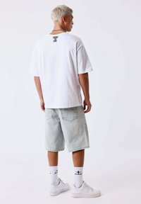 Joven con el pelo corto teñido de rubio, lleva una camiseta blanca con el lema "CHANGE THE GAME", pantalones cortos vaqueros claros, calcetines blancos y zapatillas blancas, de espaldas.