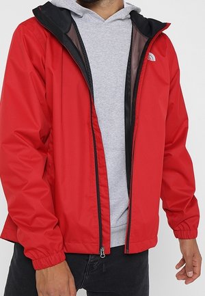 Mężczyzna w czerwonej kurtce North Face na jasnoszarej bluzie z kapturem i czarnych spodniach, z rękami swobodnie opuszczonymi wzdłuż ciała.