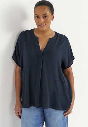 Kvinde iført en marineblå bluse med korte ærmer og V-hals samt lyseblå jeans, stående foran en ensfarvet hvid væg.