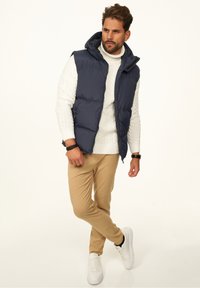 Marineblaue gesteppte Weste mit Kapuze, weißer Zopfmuster-Pullover, beige Hose und weiße Turnschuhe. Enthält einen Reißverschluss und frontale Taschen.