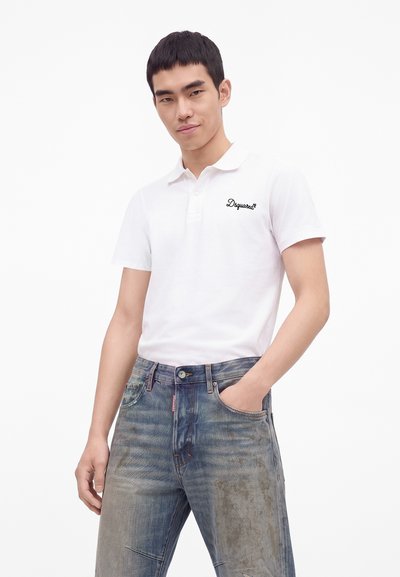 Jeune homme portant un polo blanc à manches courtes avec un logo et un jean bleu délavé, debout avec une main dans la poche sur un fond uni.