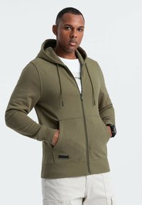 Khaki Zip-Hoodie mit Kordelzügen und Taschen. Hergestellt aus weichem Stoff, mit gerippten Bündchen und einem Etikettakzent am Saum.