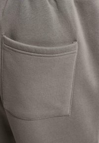 Grå bomullssweatpants med en horisontal lomme. Stoffet har en jevn tekstur og subtil stretch. Sømmen er synlig langs lommens kanter.
