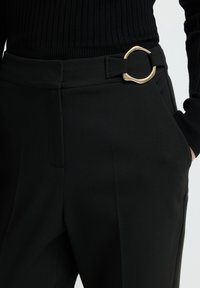 Zwarte hoge taillebroek met een gouden ronde detailsluiting, gedragen met een zwarte geribbelde longsleeve top, hand in zak.