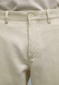 Pantalons en coton beige avec un tissage texturé, dotés d'une fermeture à bouton et de passants de ceinture. Le tissu semble durable et lisse.