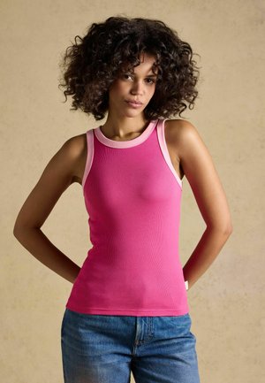 Woman with curly hair wearing a sleeveless pink top with light pink trim and blue jeans, standing with hands behind her back.