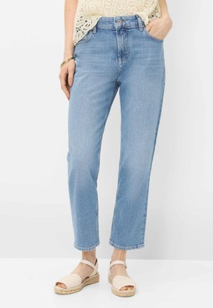 Lyseblå jeans med lige ben båret med cremefarvet hæklede top og cremefarvede espadrillesandaler, med den ene hånd i lommen, stående på hvidt gulv.