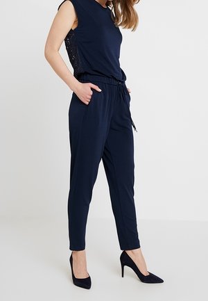 Granatowy kombinezon bez rękawów, z elastycznym pasem i kieszeniami bocznymi. Strukturalne akcenty koronkowe po bokach; zestawiony z szpiczastymi obcasami.