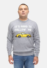 Maglione grigio lavorato a maglia con un design di un'auto gialla pixelata con il testo "È DURA SEGUIRMI" sopra. Pantaloni in denim casual.