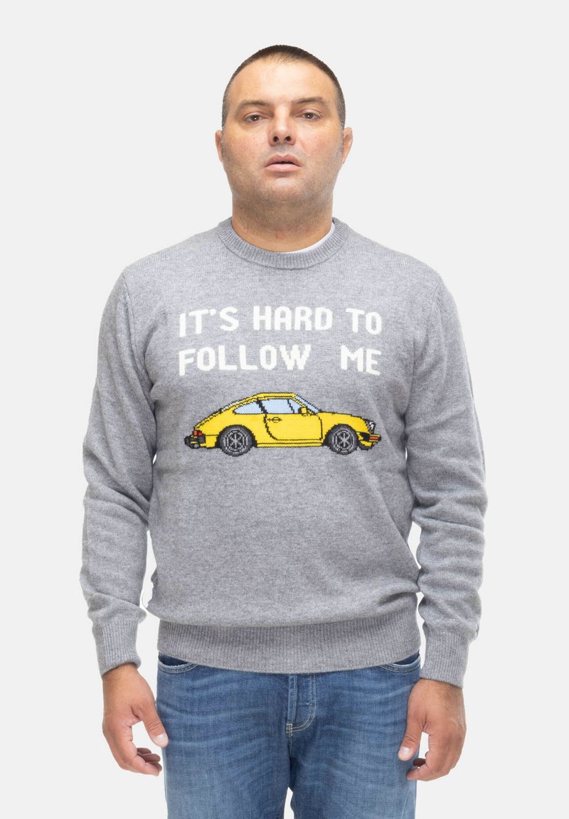 Maglione grigio lavorato a maglia con un design di un'auto gialla pixelata con il testo "È DURA SEGUIRMI" sopra. Pantaloni in denim casual.