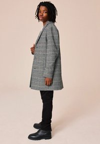 Cappotto grigio a quadri con un design strutturato, tasche laterali e maniche lunghe, abbinato a pantaloni neri e stivali di pelle nera.