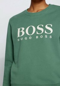 Grön sweatshirt i mjukt tyg, med ribbad halsringning och ärmar. Den har "BOSS" och "HUGO BOSS" tryckt i vitt över framsidan.