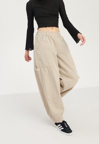Pantaloni cargo beige larghi con tasche frontali, vita con lacci e tessuto leggero. Indossati con un top nero a maniche lunghe e sneakers nere.
