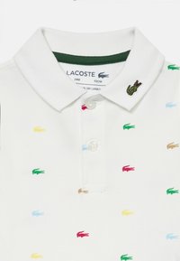 Valge polo särk, mis on valmistatud tekstiilsete puuvillast, rohelise kaelusega, väikeste punaste, siniste, roheliste, kollaste ja pruunide tikandiga krokodilli logo'dega.