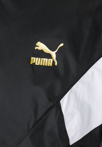 Black windbreaker featuring a white diagonal panel. Gold Puma logo and leaping cat emblem displayed prominently on the chest area.